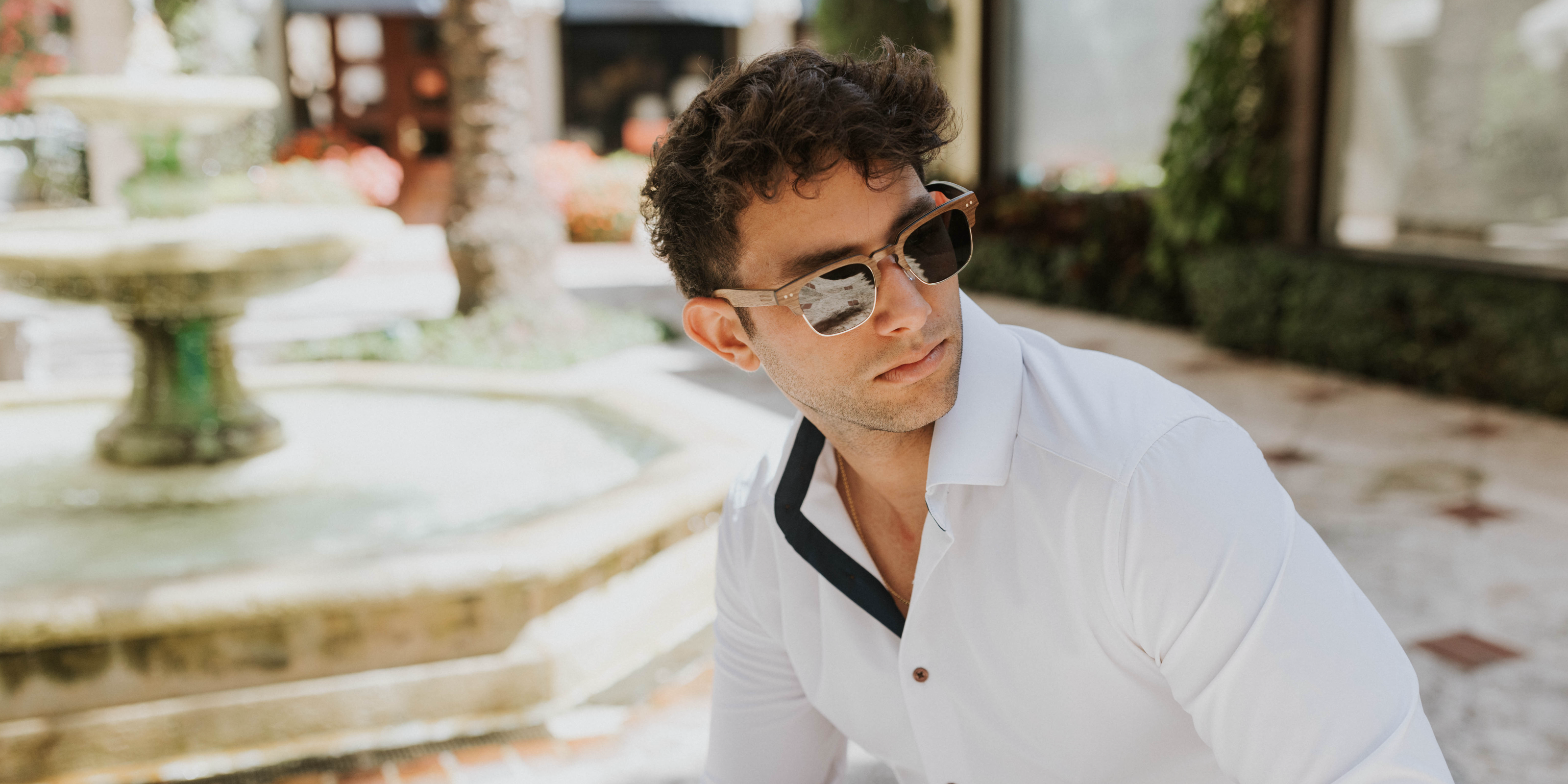 Naples walnut‑wood sunglasses worn sitting in courtyard with water fountain, three‑quarter front view.